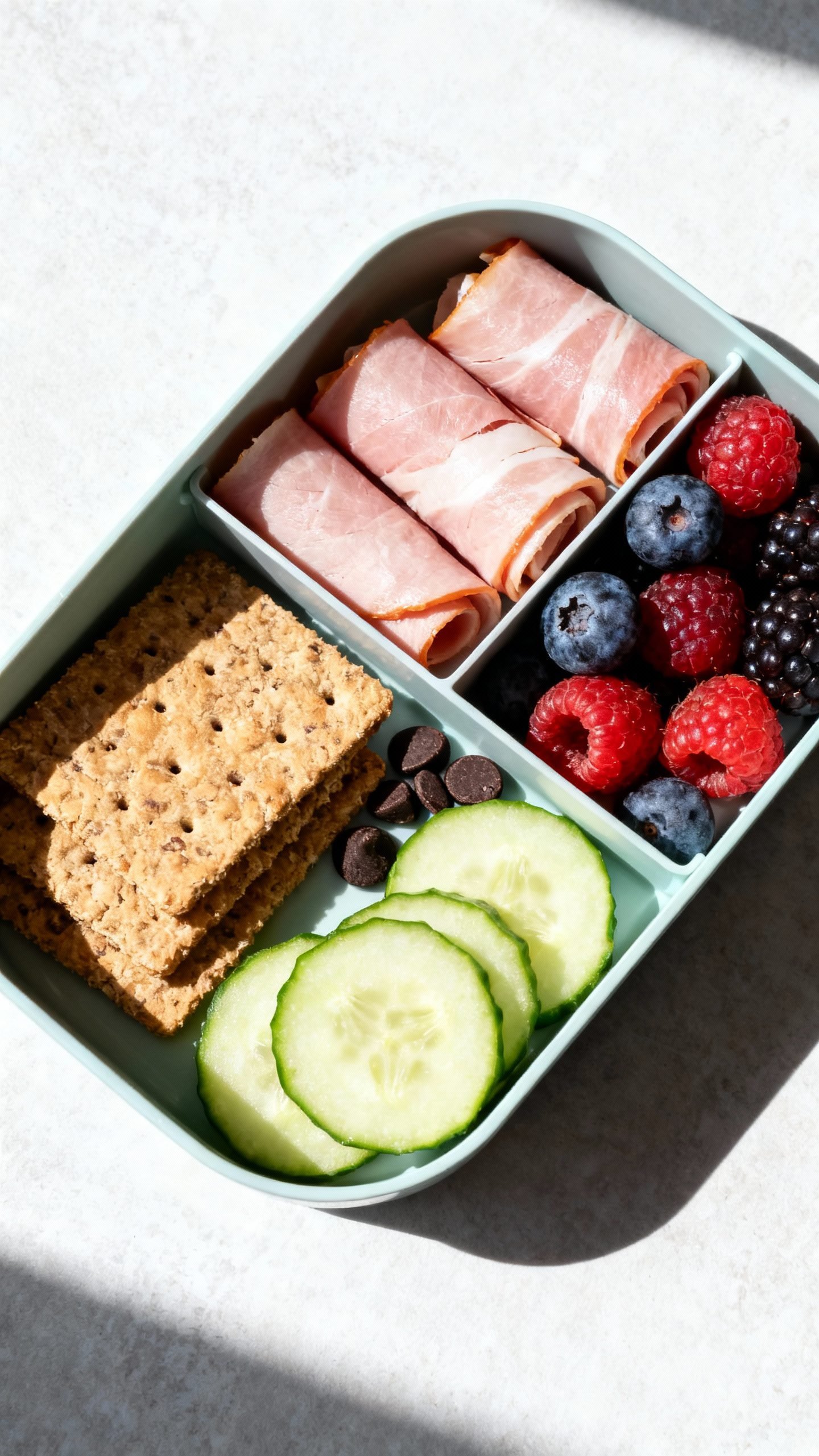 Bento box detail shot: rolled ham slices, whole-grain crackers, cucumber coins, mixed berries, few d